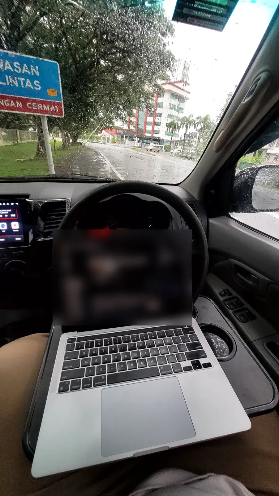 A laptop open on the steering wheel, parked under trees